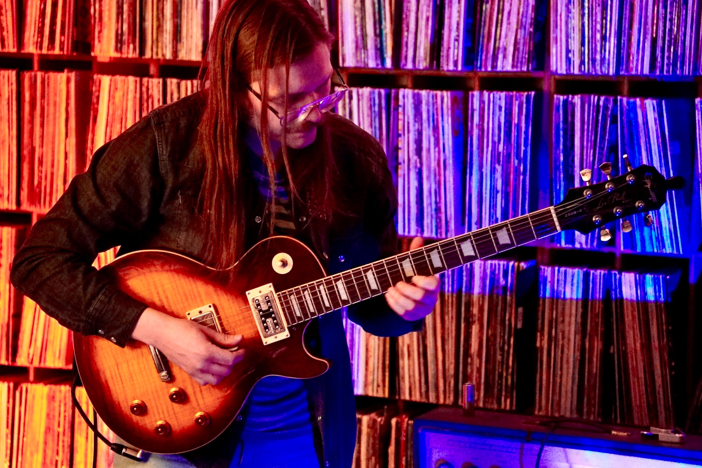 A man stands and looks at a guitar he is holding, in front of a shelf of vinyl records.
