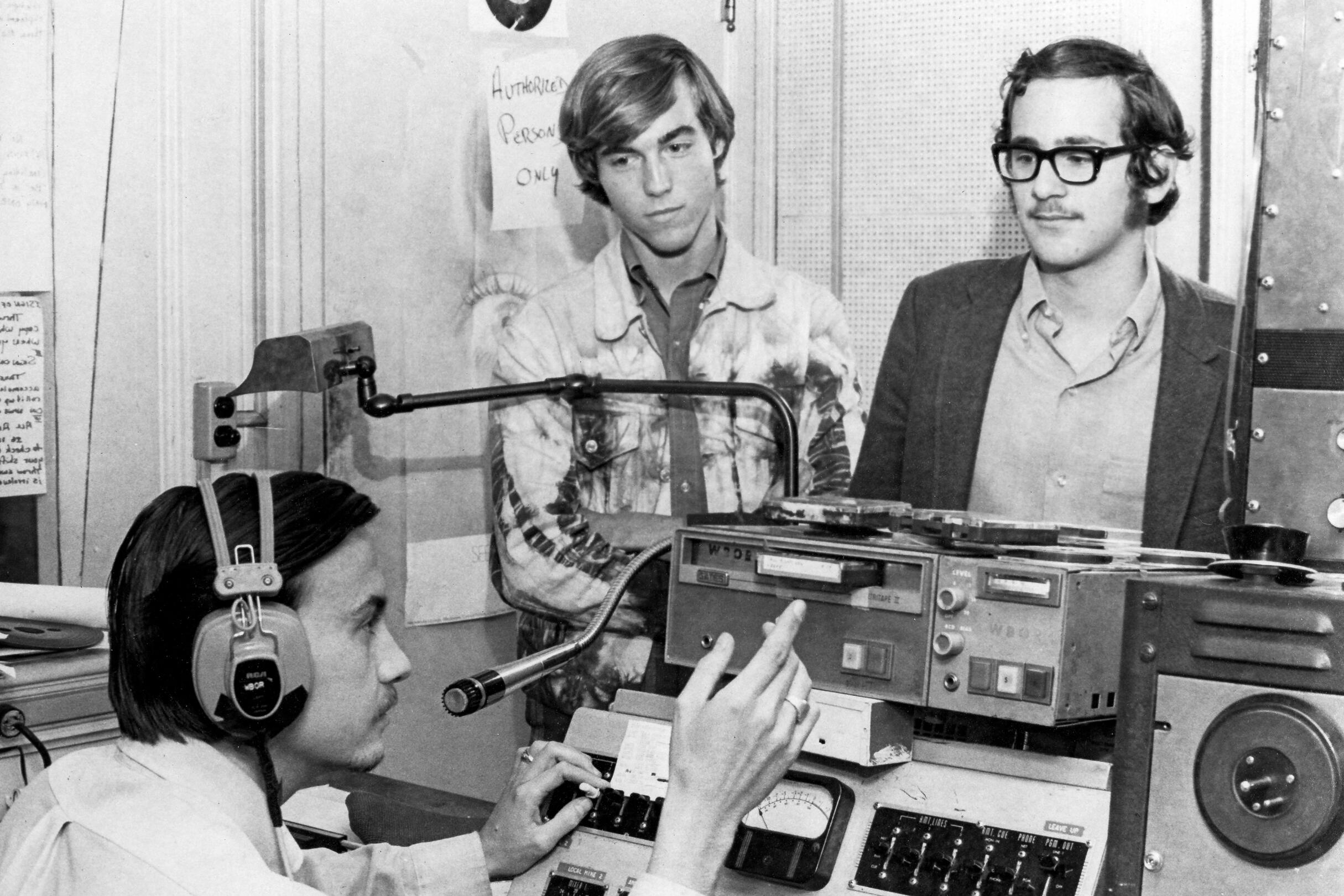 A student sits in the WBOR broadcast studio with two other students standing watching